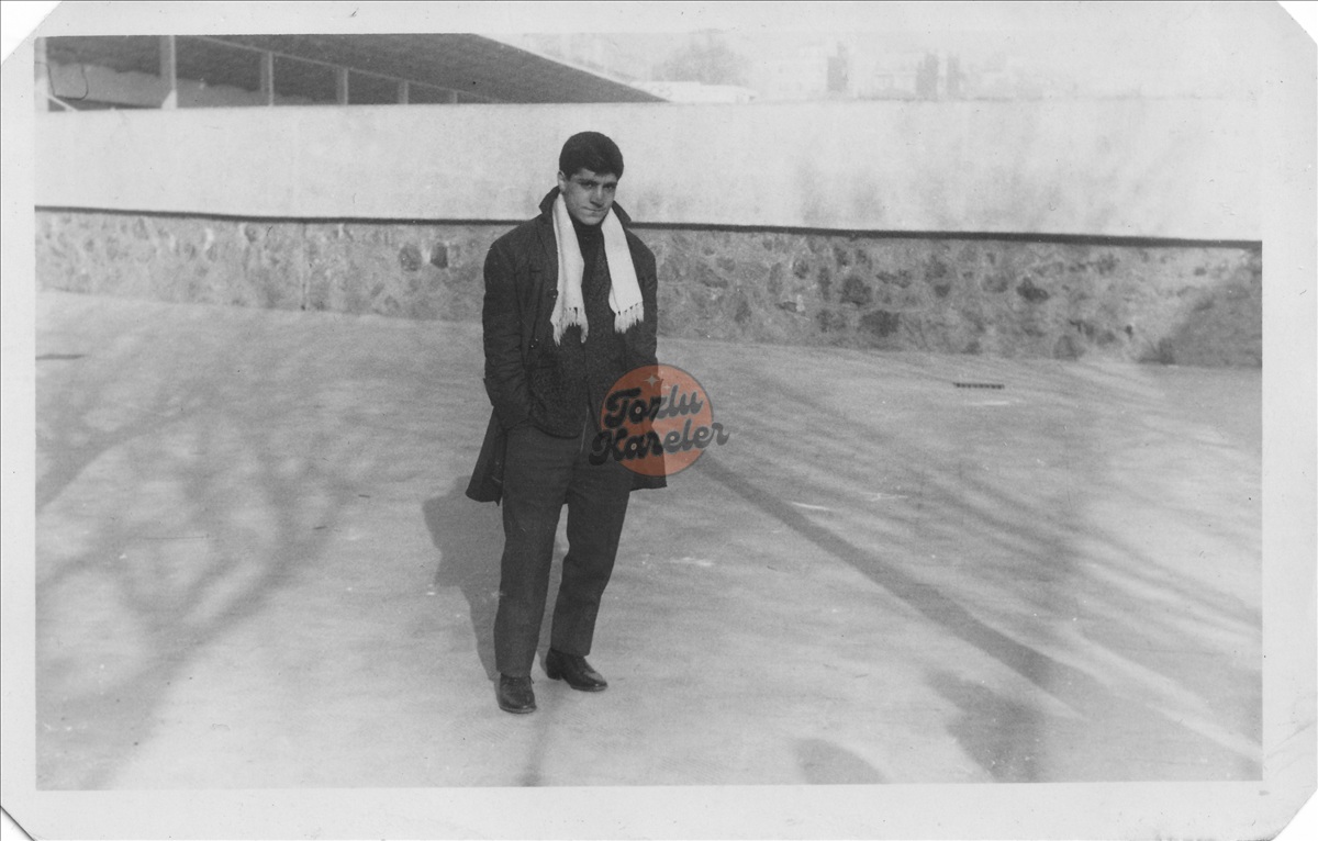 Siyah beyaz eski bir fotoğrafta; üzerinde koyu renkli palto, kumaş pantolon ve boynunda açık renkli atkı olan genç bir adam açık alanda ayakta duruyor.